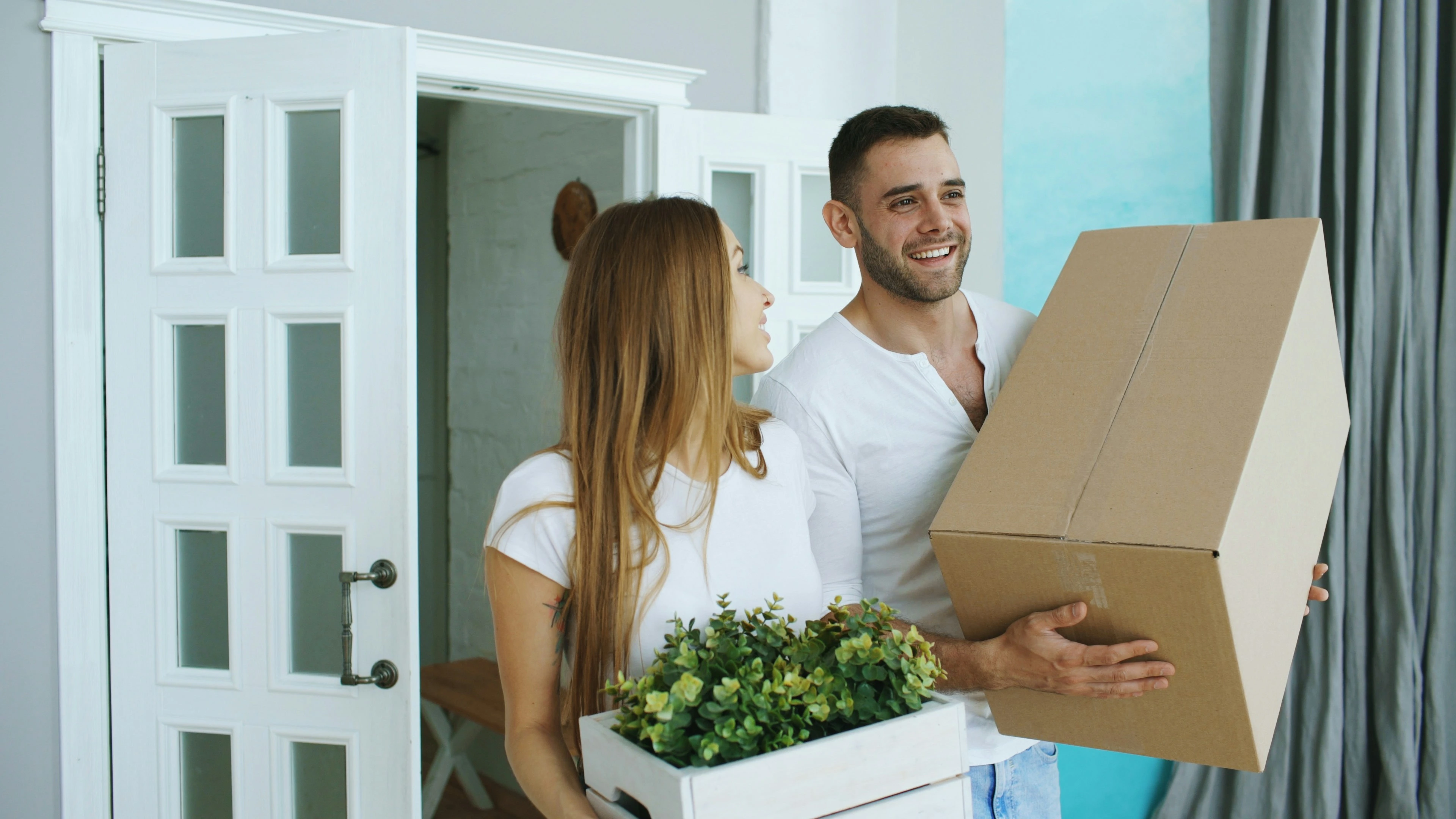 Smiling couple carrying boxes and plants while moving into a modern home, representing new homeowners in Calgary’s Altadore neighbourhood.