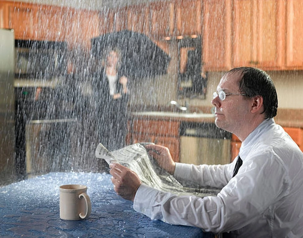 Man sitting under water pouring from a kitchen ceiling, symbolizing home inspection issues and risks for Calgary buyers.